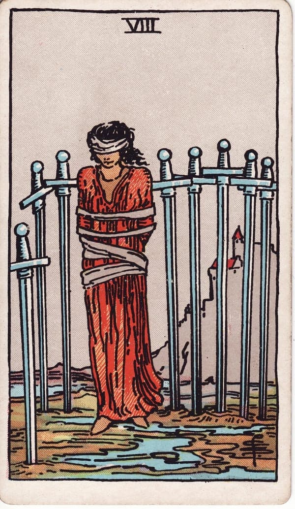 Eight of Swords tarot card: A woman stands bound and blindfolded on wet, gray ground, eight swords planted around her in an open arc — a fence with no gate because it needs none; the way ahead is simply clear.