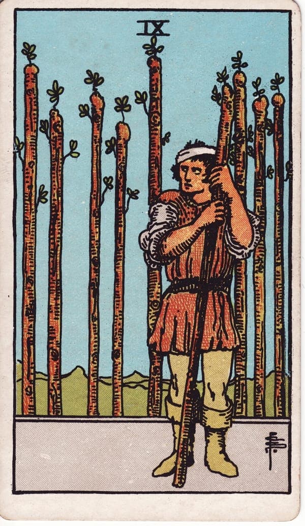 Nine of Wands tarot card: A man leans on one wand, head bandaged, eyes sidelong toward whatever might come next; eight staves stand behind him like a palisade with one gap — the gap he fills.