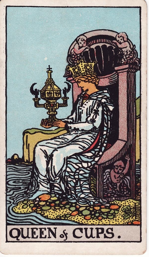 Queen of Cups tarot card: A queen sits on a throne at the ocean's edge, decorated with sea-nymphs and shells, gazing intently at an ornate covered cup with angel-winged handles — the only lidded cup in the deck.