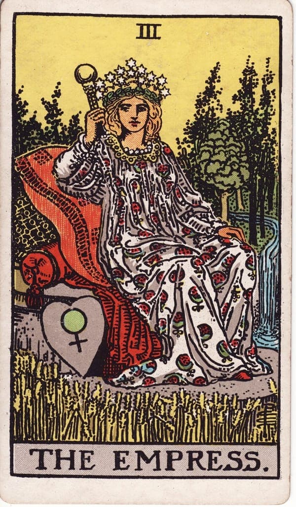 The Empress tarot card — A crowned figure reclines on cushioned luxury in the middle of a ripening wheat field, a river windi