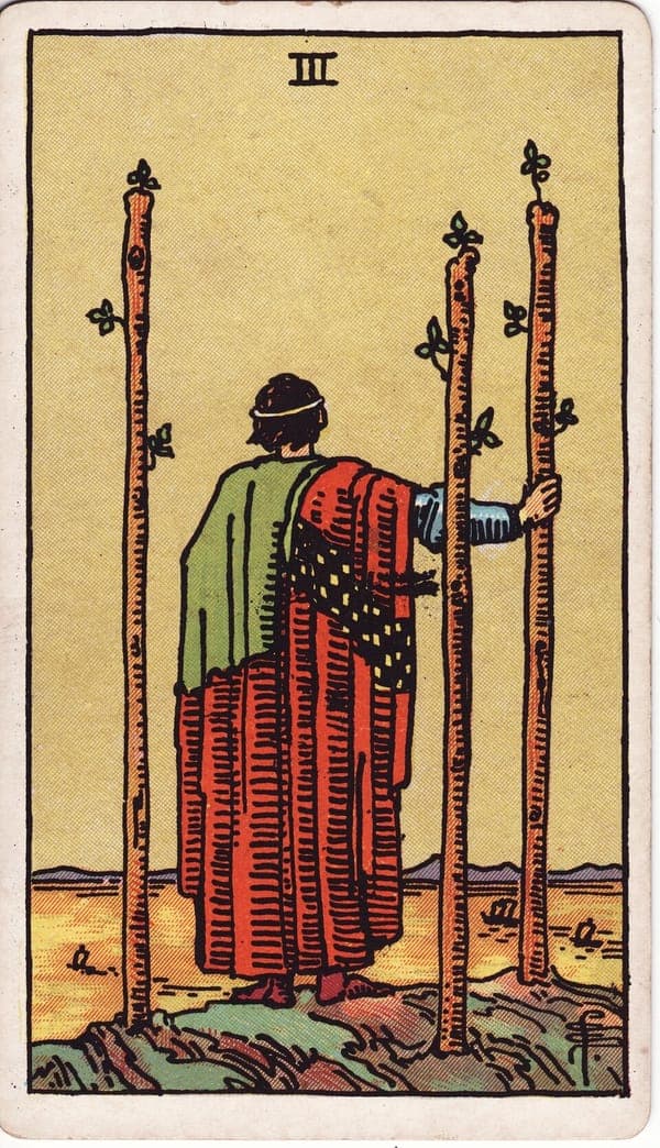 Three of Wands tarot card: A robed figure stands on a cliff's edge, back to the viewer, gazing over a golden sea where ships move in the distance.