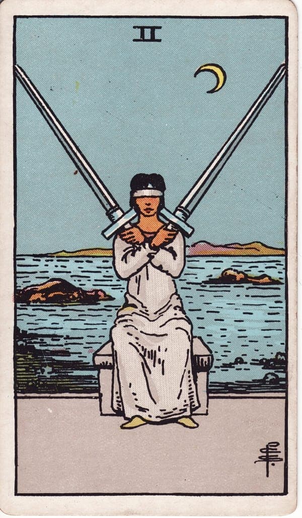 Two of Swords tarot card: A blindfolded woman in gray sits on a stone bench before the sea, two long swords crossed over her chest in perfect balance.