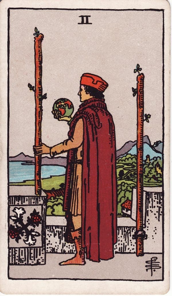 Two of Wands tarot card: A figure stands on a castle battlement, a small globe held in one hand and a planted wand in the other — the world literally in hand, the second wand bolted to the wall behind.