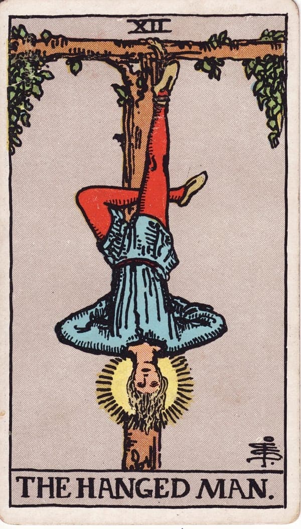 The Hanged Man tarot card — A man hangs by one foot from a living T-shaped tree, his free leg crossed behind, arms folded — his